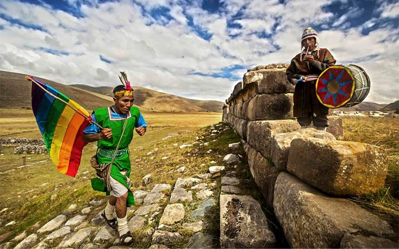 A modern day Chacquis runner in the Andes, carrying a rainbow flag running up a stone path toward an ancient wall where another man in Quechuan clothing is playing a large, multicolored drum.