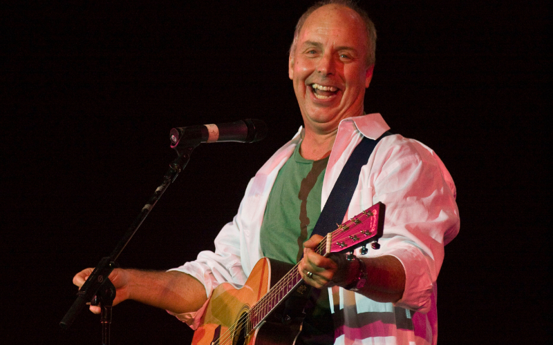 Comedian, musician and all around nice guy, Bill Harley smiling at the camera while in the spotlight on a dark stage, holding a guitar, in front of a microphone.