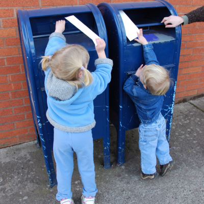 Two children reaching up to put envelopes into two USPS postal boxes.