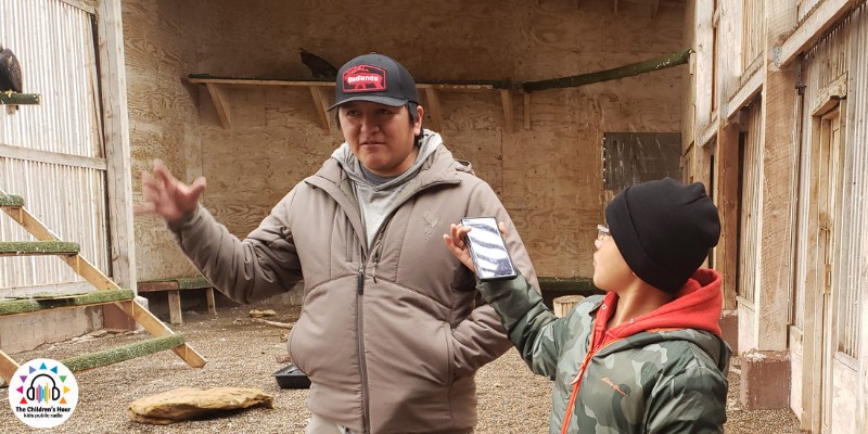 A child wearing a black hat, orange hoodie with a green overcoat, is holding up a phone recorder to the Zuni Eagle Aviator caretaker, who is wearing a blue cap with orange lettering, a tan coat, and gesturing with his right hand in an open palm stretched out a bit to the side. There is a little bit of an eagle visible in the background but only its back and tailfeathers can be seen. The Children's Hour logo is in the bottom left corner. The scene is inside the eagle aviary, made of wooden walls, and slatted sides, with perches made of bark stripped tree limbs.