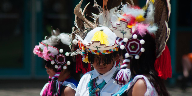 An image depicting Zuni youth dressed in traditional dance gear, including elaborately beaded and feathered head coverings, with black fringe hanging down on the face obscuring the eyes of the children. The outfits are colorful and the kids are not looking at the camera. These are Zuni eagle dancers.