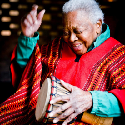 Smiling Ella Jenkins wearing a red sarape, playing a talking drum. Her hand is raised in mid-strike.