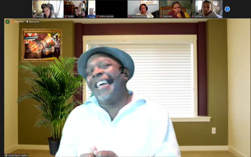 A screenshot of a Zoom screen with children at top and mostly featuring the face of Devin Walker, laughing and wearing a grey hat with a white collared shirt, sitting in front of a background screen made to look like a simple room.