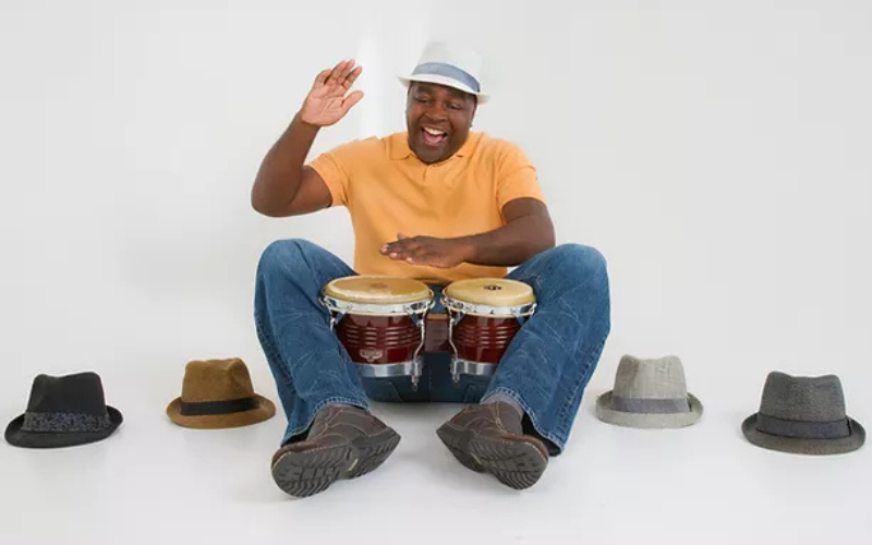 Musician Devin Walker sitting on a floor in a white hat with a black brim, surrounded by other hats and holding bongo drums between his legs. He is wearing a yellow shirt and blue jeans, and is seated with a white background.