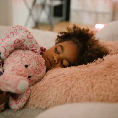 A black child clutching a stuffed animal elephant, laying asleep on a fuzzy pink pillow.