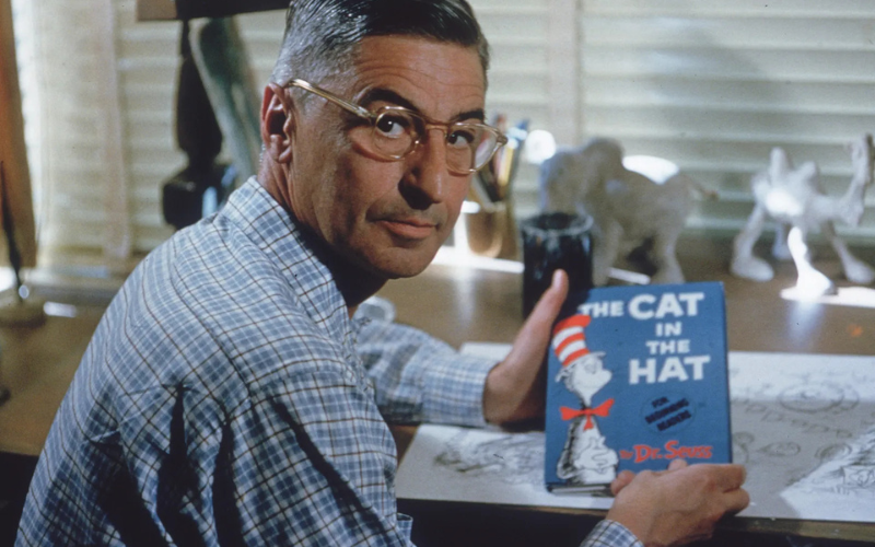 Theodor Geisel, also known as Dr. Seuss, at his desk holding a copy of Cat in the Hat.