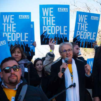 Several people standing holding signs that read "We the People ACLU" behind a woman with short grey hair, a white scarf and black coat, speaking into a microphone. 