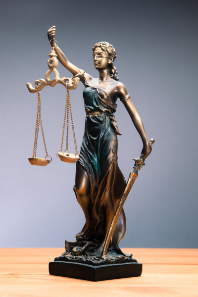 The Lady of Justice holds a sward in her left hand, scales in her right hand, with loose hair decorated with pearls, and a blindfold over her eyes. She is standing on snakes. This is a statue of the Lady of Justice which is on a wooden table surface with a grey background.