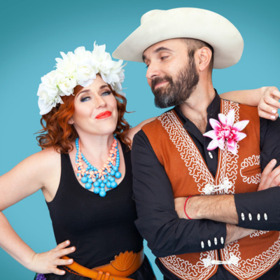 Lucky Diaz dressed in a white cowboy hat and brown vest with a dahlia pinned to its lapel, and wearing a black buttonup shirt underneath, is looking at Alicia Gaddis. She is wearing a hat of white flowers, a black tank top, and a blue turquoise necklace, as well as a brown belt. Alicia is looking straight into the camera with a closed-mouth smile.